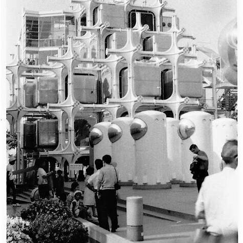 Fotografía en blanco y negro de un edificio experimental con módulos cúbicos y una estructura blanca enrejada. En la parte inferior, se observa a un grupo de personas explorando la exhibición. Diseño icónico del siglo XX con estética retrofuturista y arquitectura metabolista