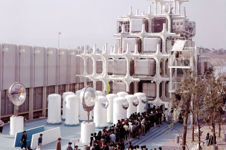 Fotografía tomada desde un ángulo amplio, mostrando una larga fila de visitantes esperando para entrar a un pabellón arquitectónico innovador. La estructura presenta módulos cúbicos dentro de un entramado blanco y elementos cilíndricos con esferas reflectantes.