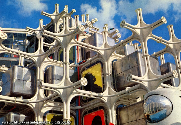 Fotografía a color de un edificio experimental con una estructura modular compuesta por cubos metálicos insertados en un entramado blanco con conexiones orgánicas. Destacan ventanas con paneles de colores y elementos cilíndricos reflectantes en la parte inferior. El cielo azul con nubes refuerza el contraste visual del diseño vanguardista.